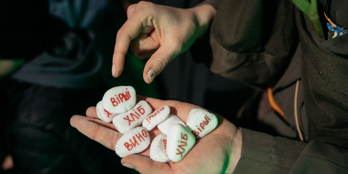 І в жмені гладенькі білі камінці, на яких виведено червоним «Вірші», «Хліб», «Вино».webp